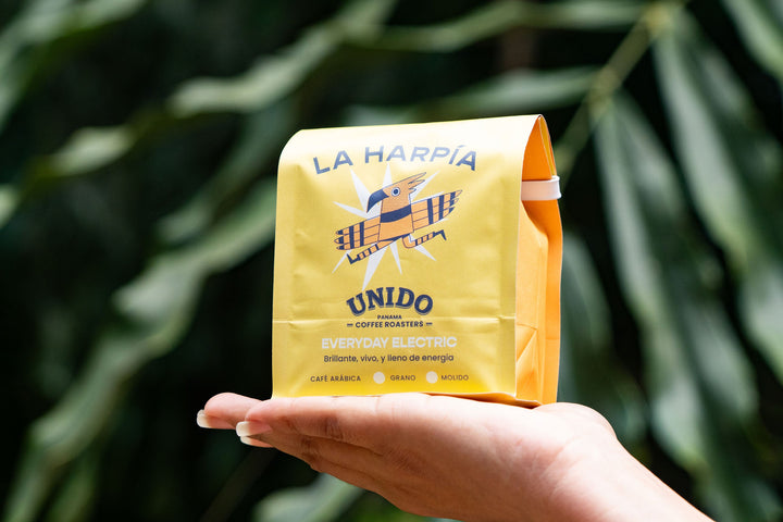 A hand holding a yellow bag of La Harpía Unido coffee roasters, with a blurred green leafy background.